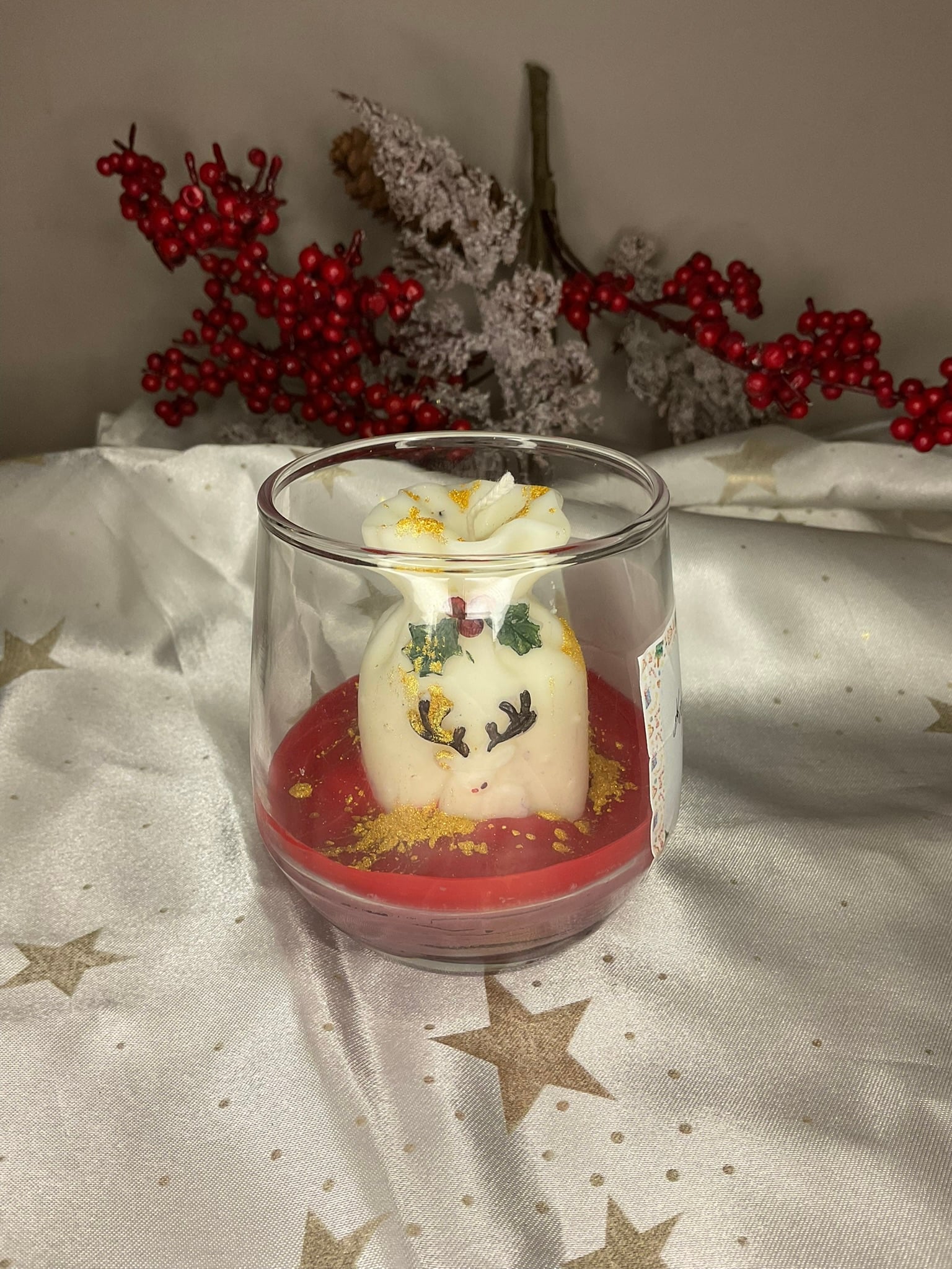 Χριστουγεννιάτικο κερί Winter Spark Gift Bag σε γυάλινο ποτήρι με λευκό σάκο δώρων, ζωγραφισμένο τάρανδο, κόκκινη βάση και χρυσόσκονη.